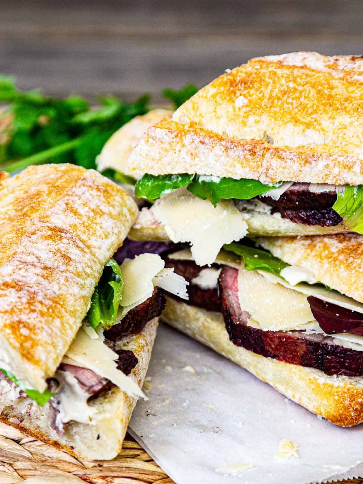 Three stacked and sliced roast beef sandwiches on French bread with lettuce, cheese and a creamy spread on parchment paper ready to eat.