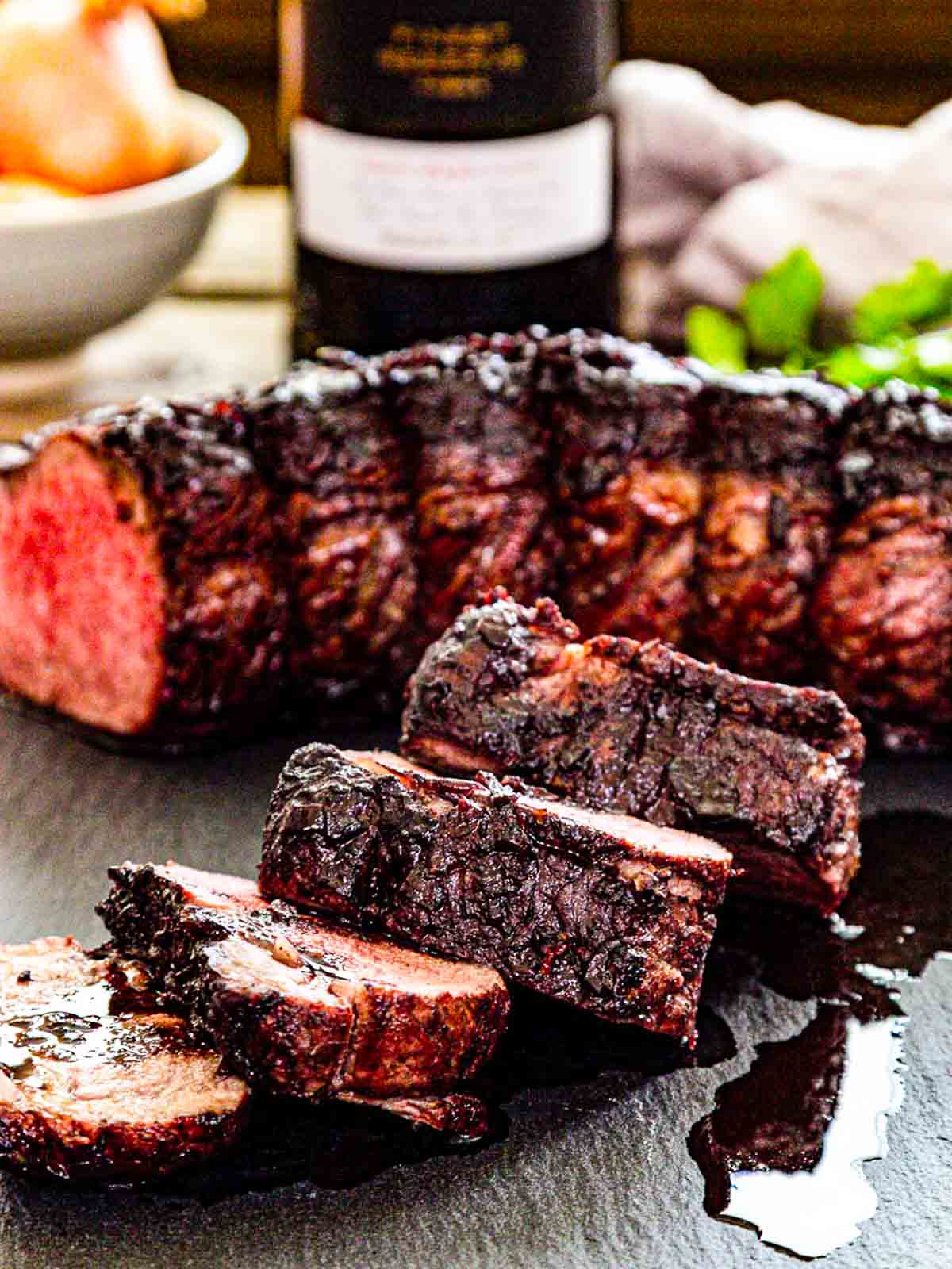 Four thick slices of roast beef with the whole roast in the background and juices running down the black slate cutting board.