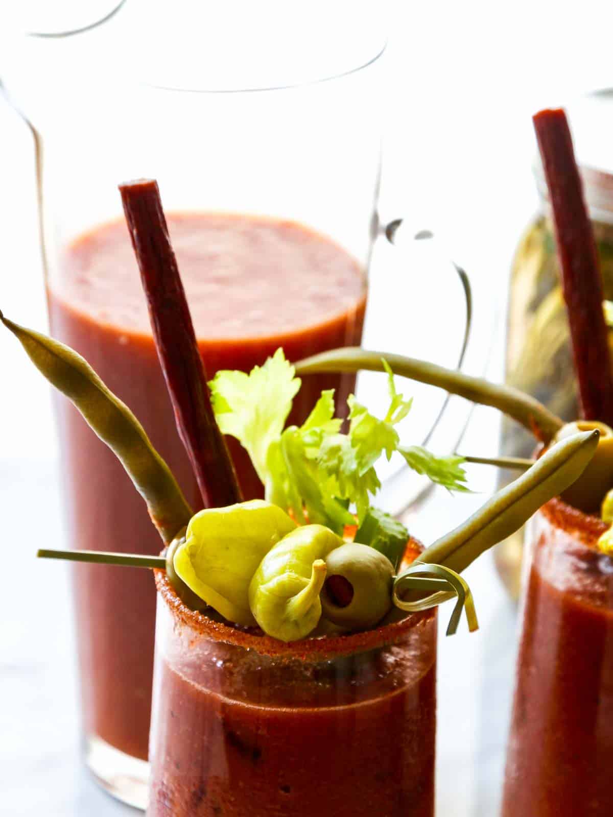A pitcher of Bloody Mary mix with two glasses garnished with olives, pepperoncinis, and pepperoni sticks.