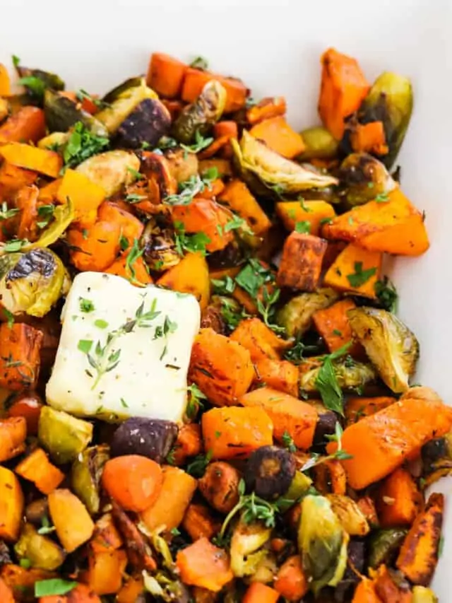 A square white dish filled with colorful roasted vegetables and topped with squares of butter garnished with herbs.