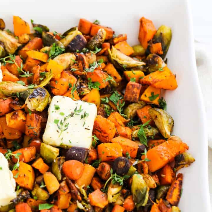 A square white dish filled with colorful roasted vegetables and topped with squares of butter garnished with herbs.