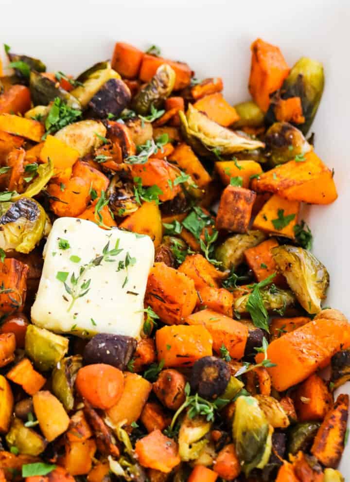 A square white dish filled with colorful roasted vegetables and topped with squares of butter garnished with herbs.