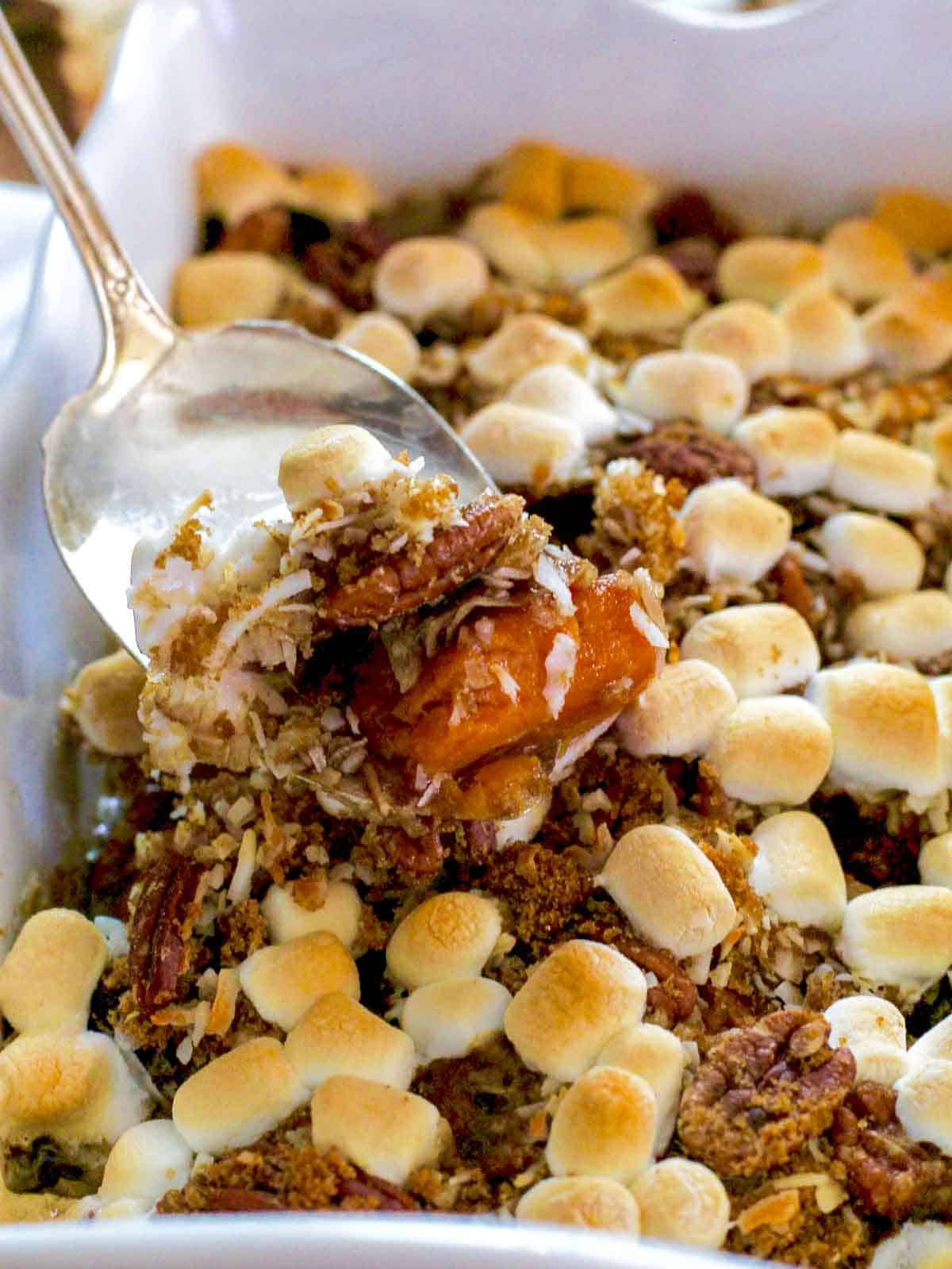 A silver server scooping up sweet potato casserole with golden toasted marshmallows on top out of a white casserole dish.