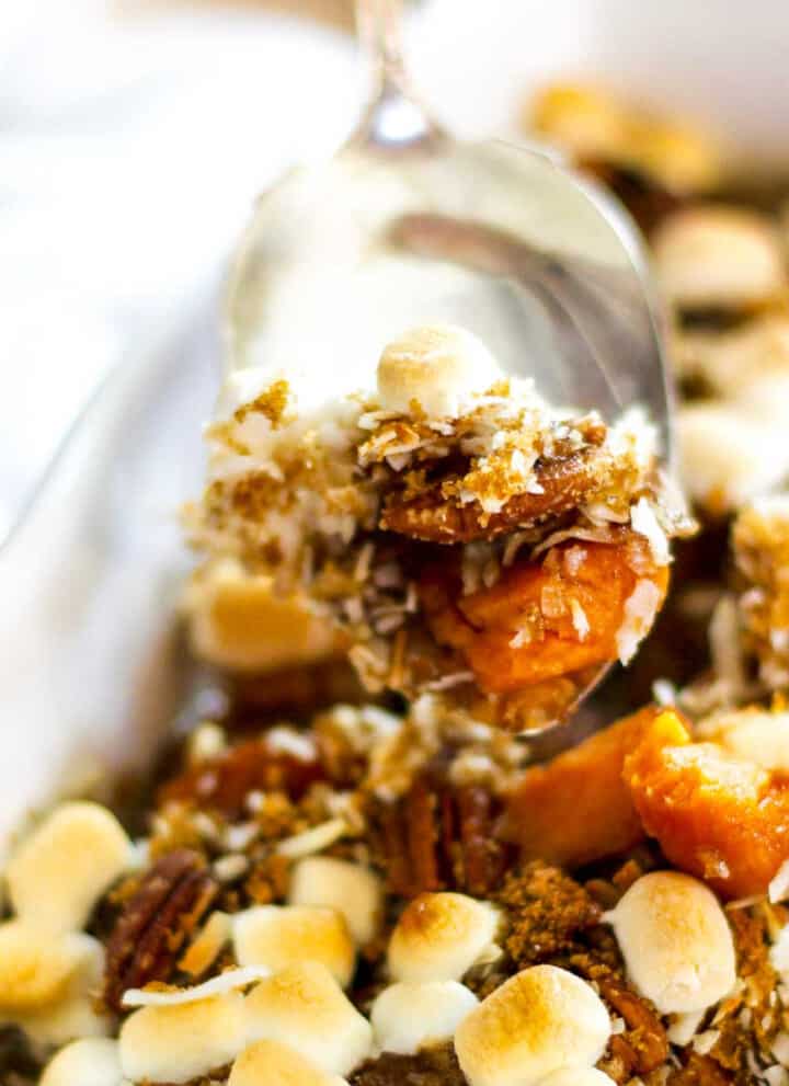 A silver server scooping up sweet potato casserole with golden toasted marshmallows on top out of a white casserole dish.