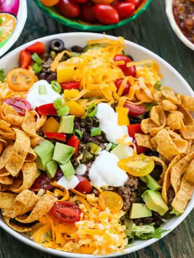 A white plate full of taco salad topped with taco meat, grated cheese, Fritos, sour cream, black olives and tomatoes.