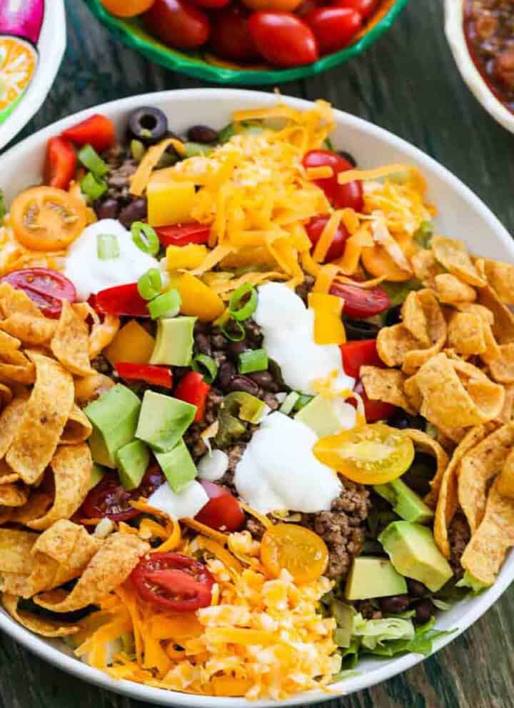 A white plate full of taco salad topped with taco meat, grated cheese, Fritos, sour cream, black olives and tomatoes.