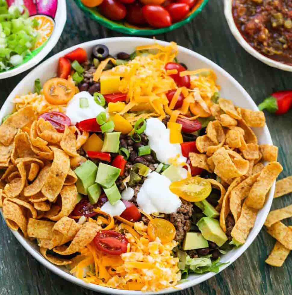 A white plate full of taco salad topped with taco meat, grated cheese, Fritos, sour cream, black olives and tomatoes.