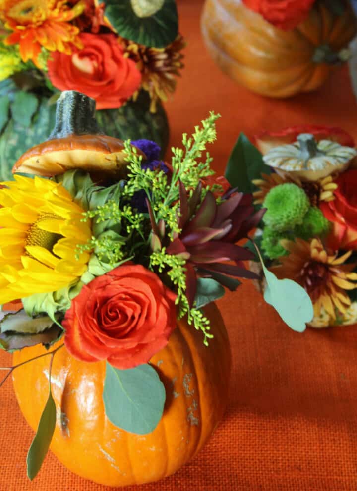 An orange table runner with handmade orange pumpkins filled with fresh Fall flowers and greenery for Thanksgiving centerpieces.