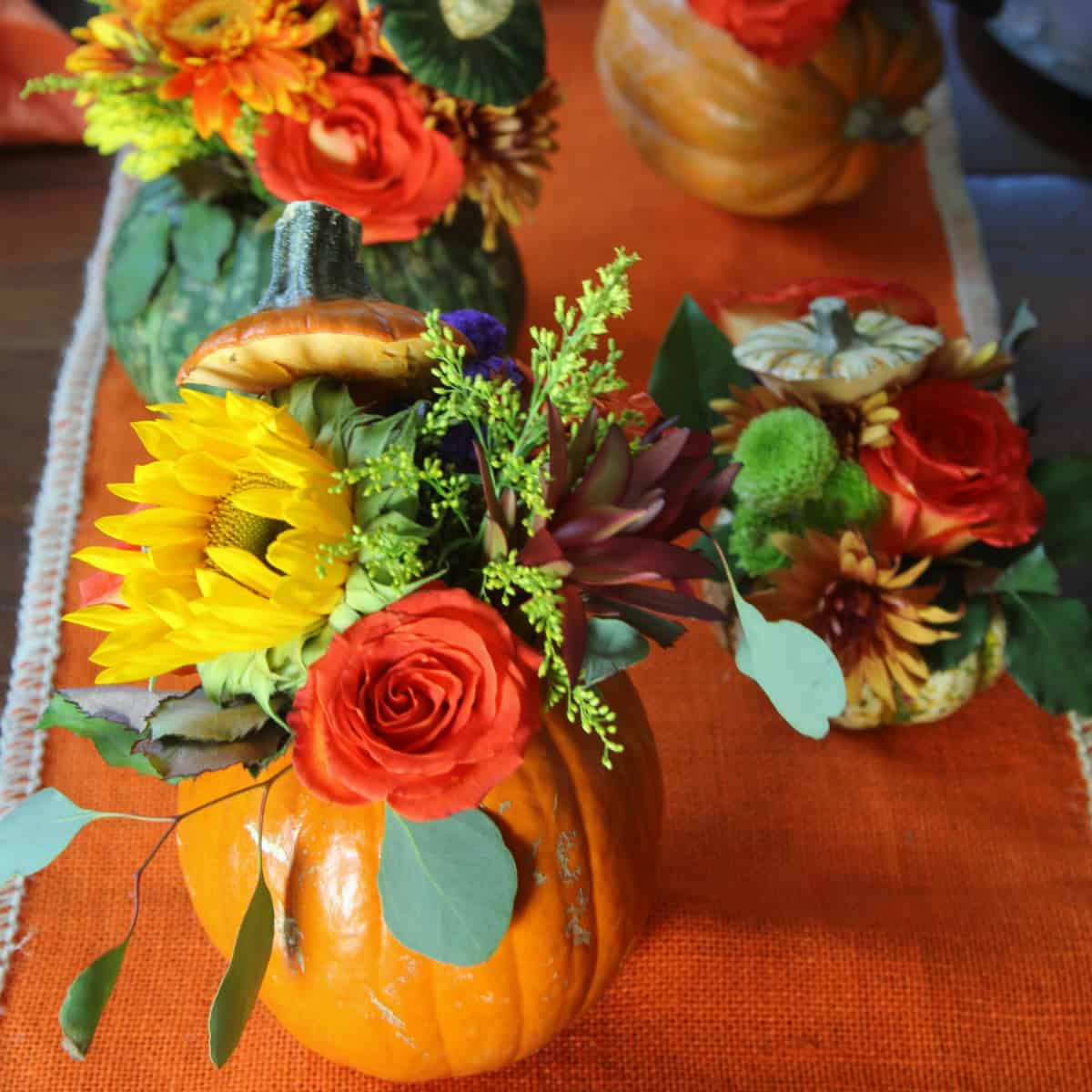 An orange table runner with handmade orange pumpkins filled with fresh Fall flowers and greenery for Thanksgiving centerpieces.