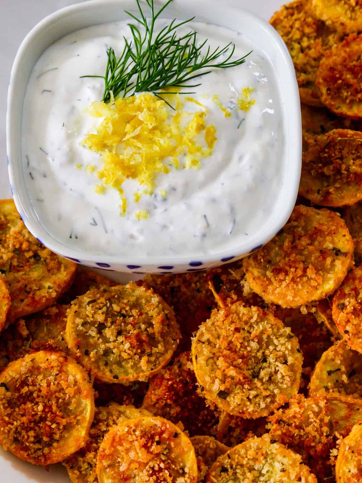 Looking down on a plate of zucchini chips with a yogurt dip garnished with fresh dill and lemon zest.