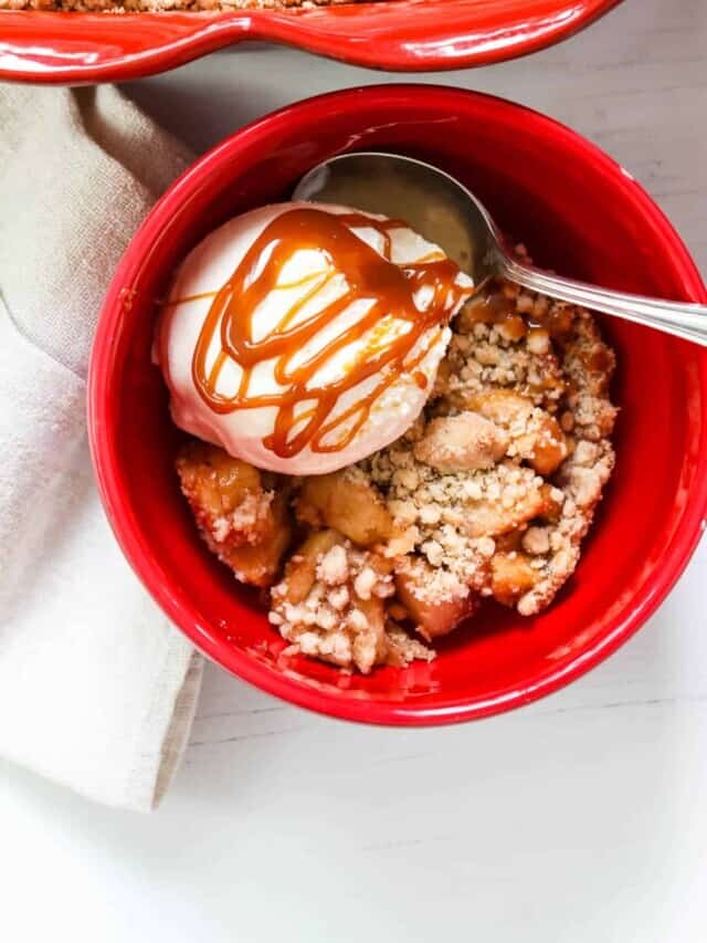 A red bowl filled with a slice of apple crisp, a scoop of vanilla ice cream, and caramel sauce.
