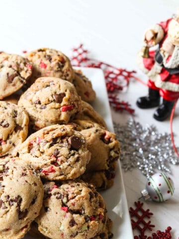 A white rectangle plate filled with chocolate chip peppermint cookies with a small Santa nearby.