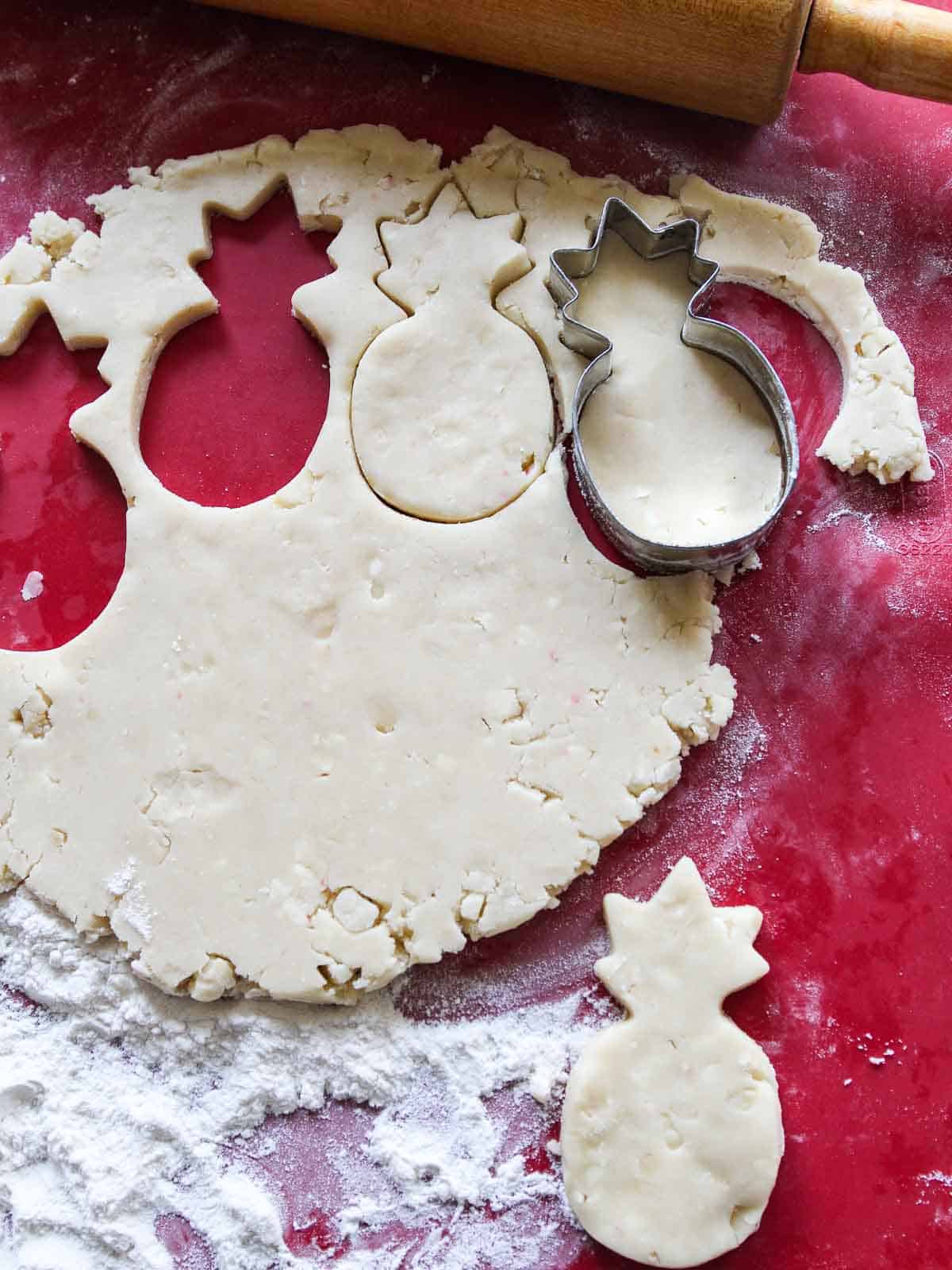 A pineapple shaped cookie cutter on a red cutting board making cookies.