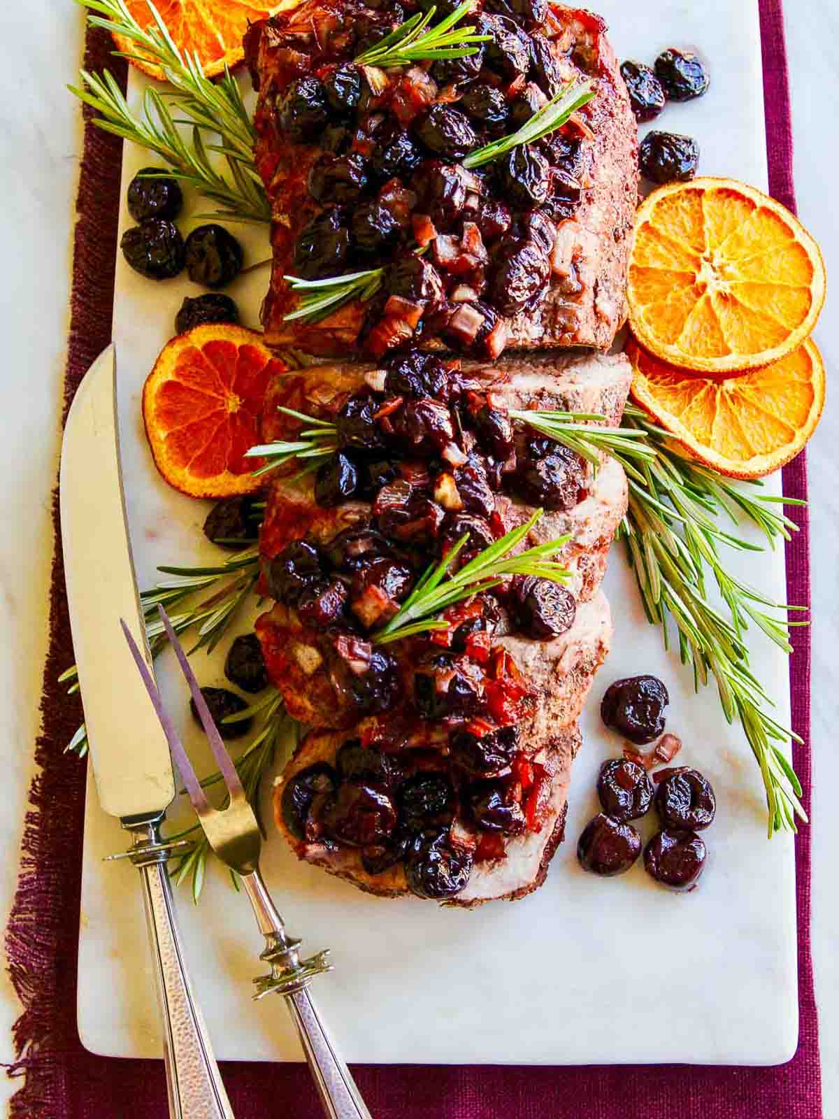 A cooked pork loin loaded with cherries and garnished with rosemary and dried oranges.