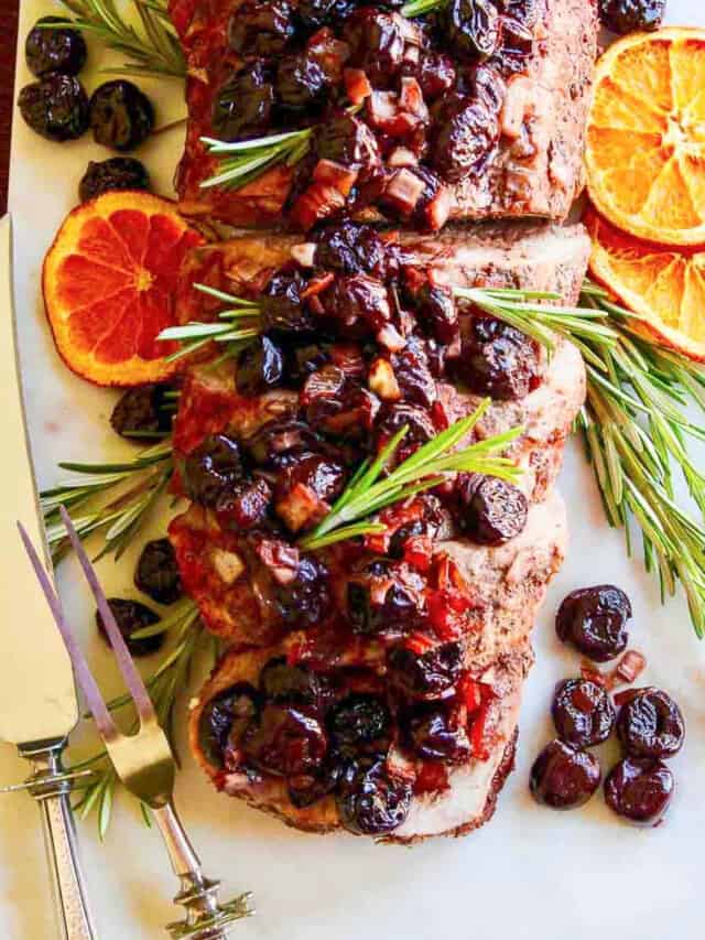 A sliver carving fork and knife next to a pork loin roast and topped with cherries.
