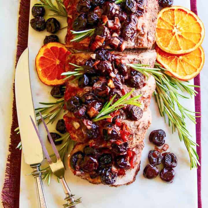 A sliver carving fork and knife next to a pork loin roast and topped with cherries.