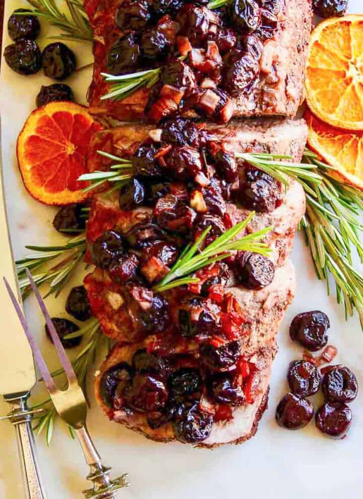A sliver carving fork and knife next to a pork loin roast and topped with cherries.