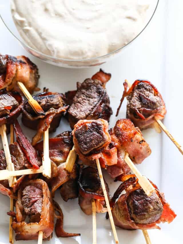 A glass bowl filled with horseradish dipping sauce with bacon wrapped filet mignon appetizers on toothpicks.