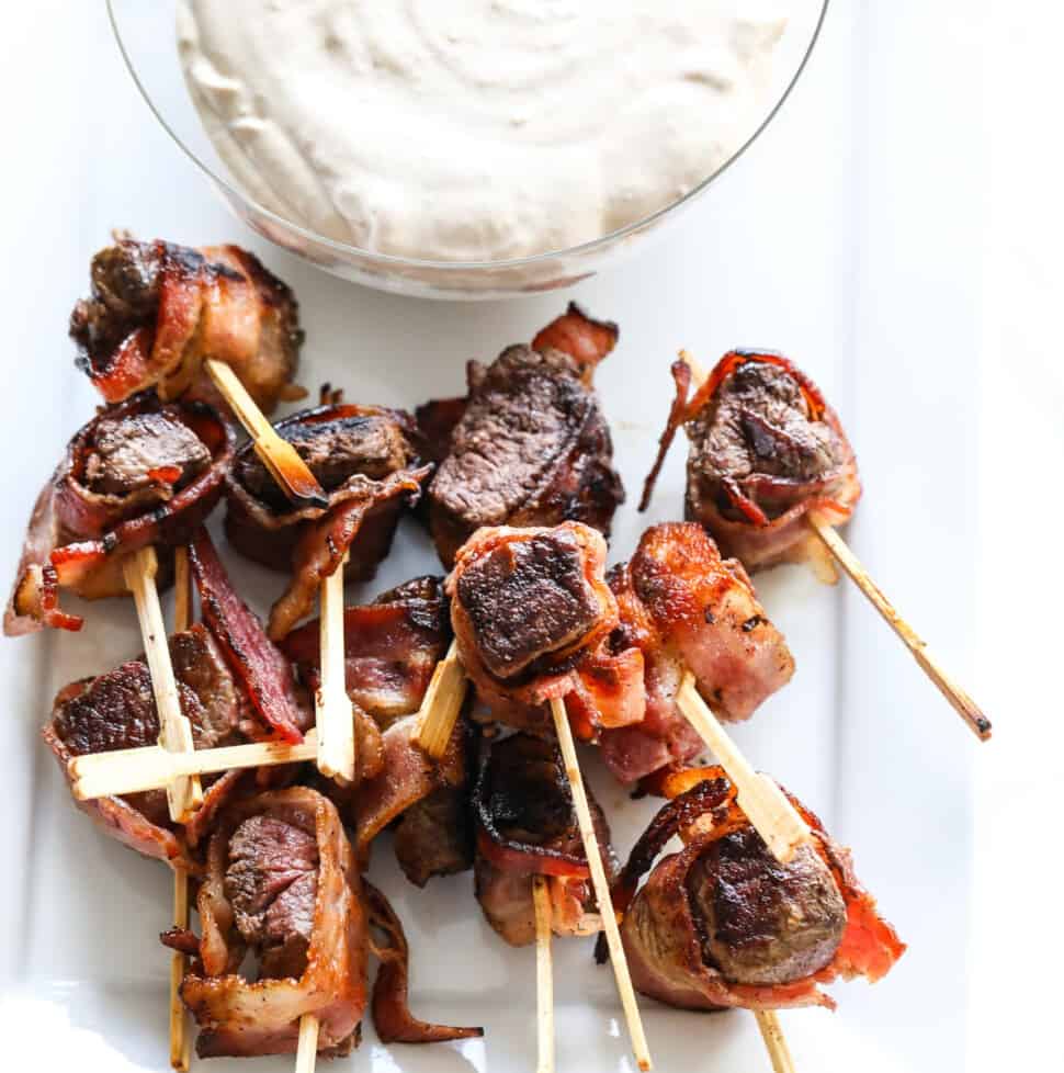 A glass bowl filled with horseradish dipping sauce with bacon wrapped filet mignon appetizers on toothpicks.