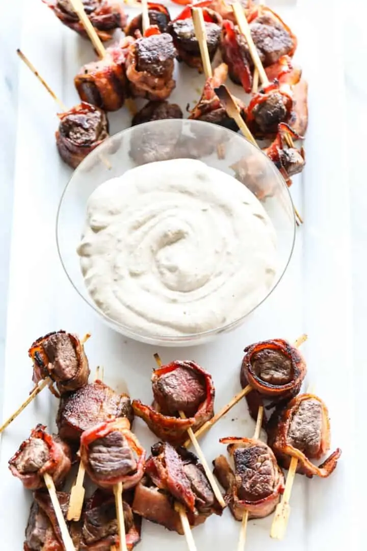 Looking down on a white platter filled with bacon wrapped filet mignon appetizers and a bowl of white horseradish sauce.