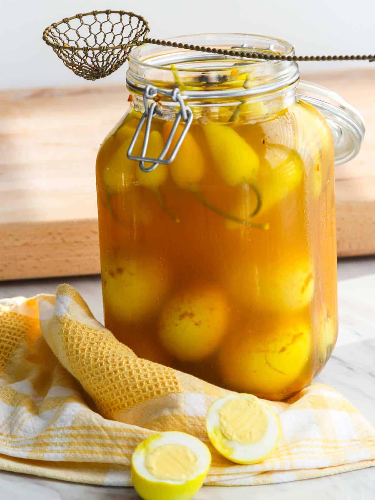 A large 2 quart clamp jar filled with yellow pickled eggs and chili peppers on a yellow and white kitchen towel.