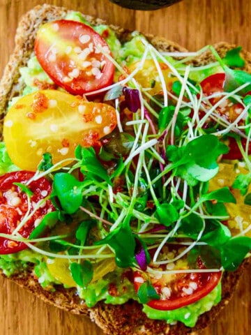 A close up photo of a slice of avocado toast topped with small heirloom tomatoes and microgreens.