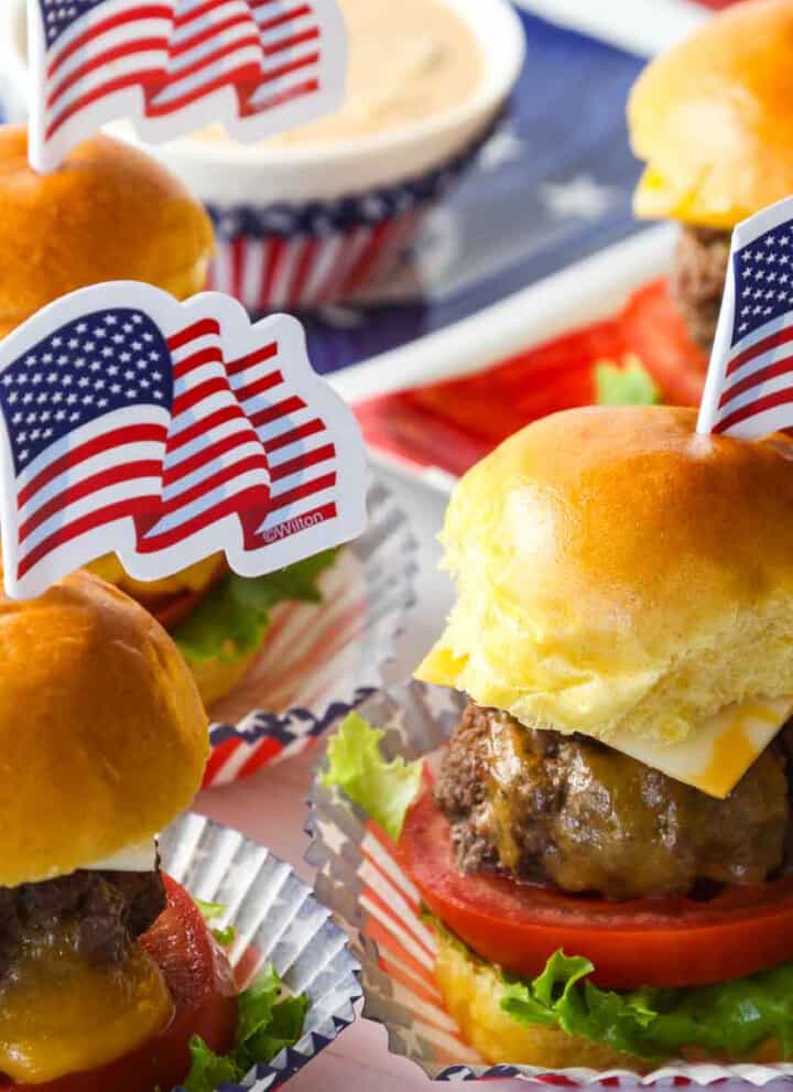 Mini cheeseburgers with melted cheese, tomato, and lettuce with American flag toothpick decoration on top.