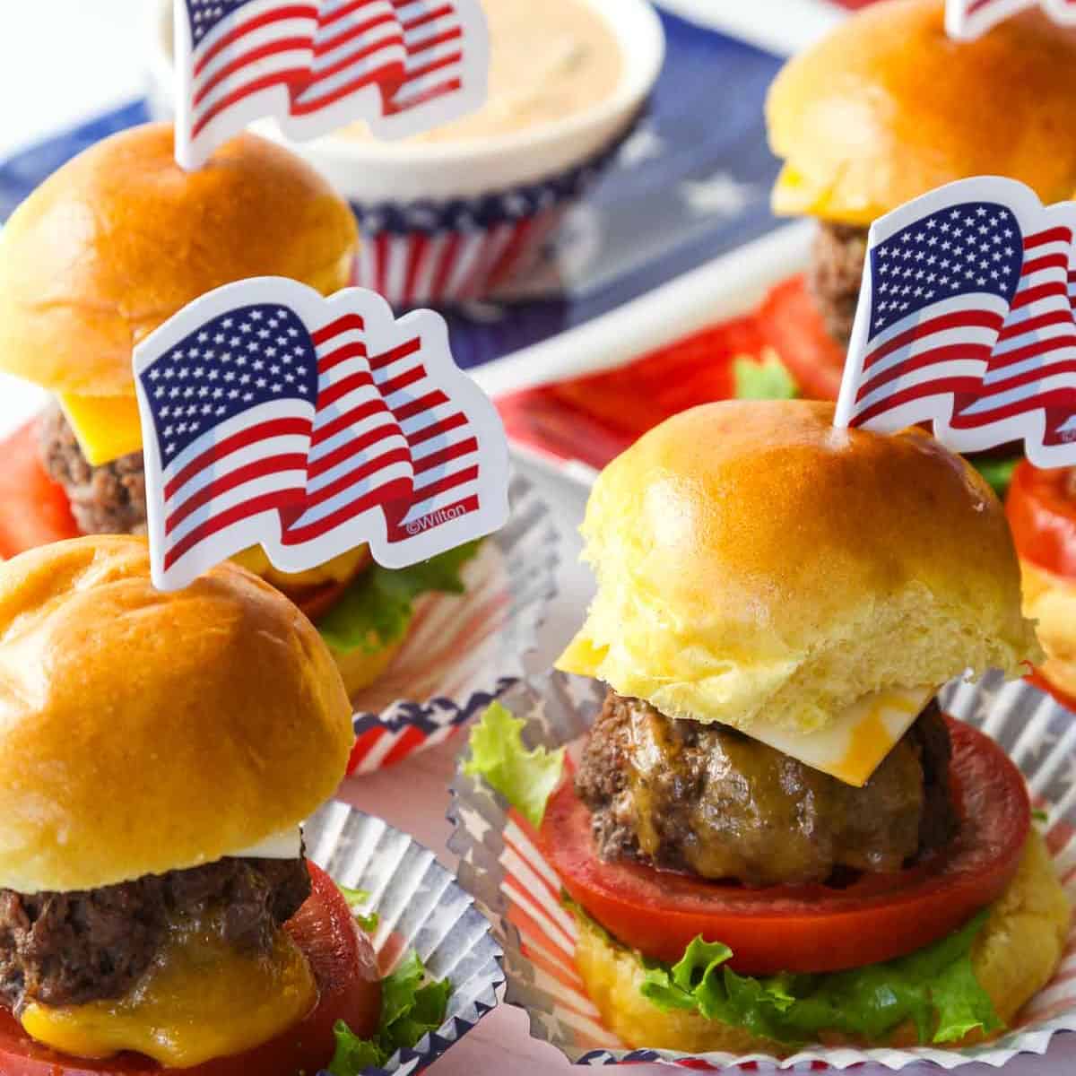 Mini cheeseburgers with melted cheese, tomato, and lettuce with American flag toothpick decoration on top.