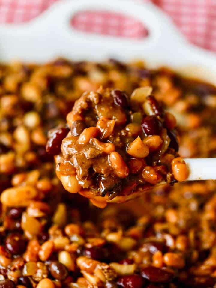 A large dish of cowboy beans with white spoon scooping out a serving.