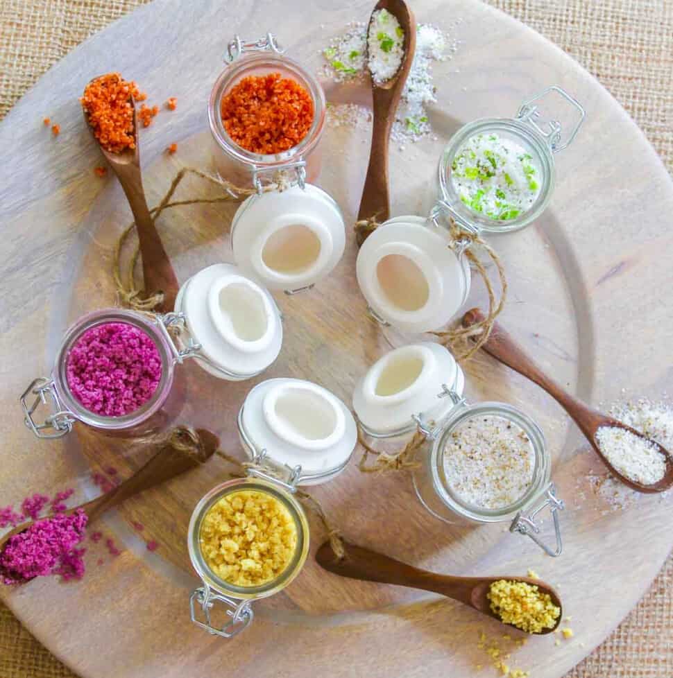 A wood platter with 5 seasoning jars open filled with flavored finishing salts.