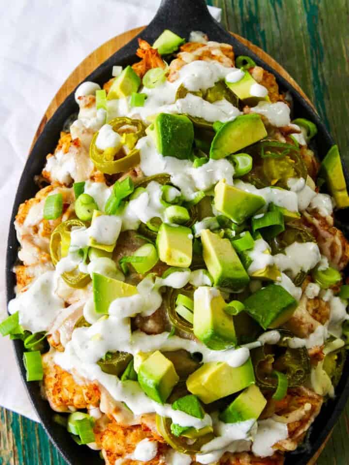 An oval cast iron pan with tater tot nachos (aka Totchos) topped with avocado, creamy sauce, and green onions.