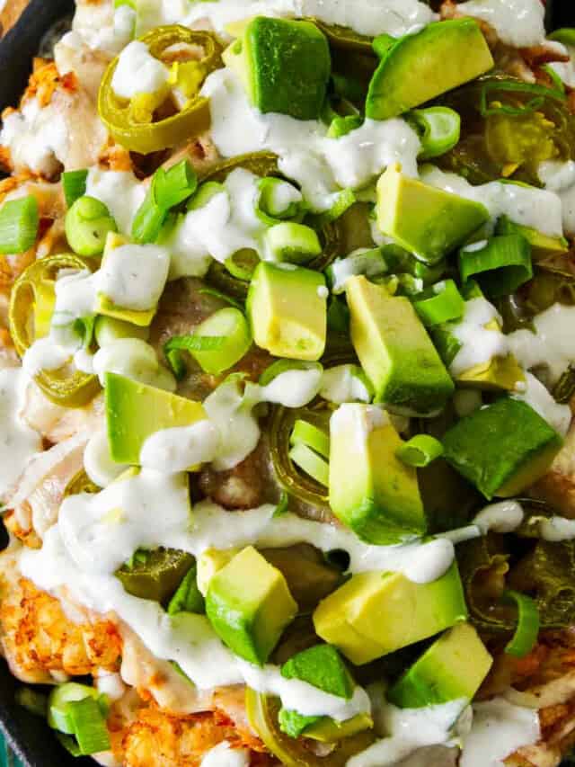 A close up of a oval cast iron pan with tater tots, pulled pork, cut avocado, and drizzled with white sauce.