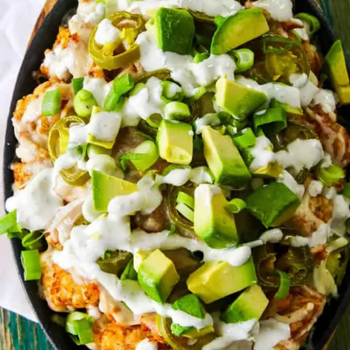 A close up of a oval cast iron pan with tater tots, pulled pork, cut avocado, and drizzled with white sauce.