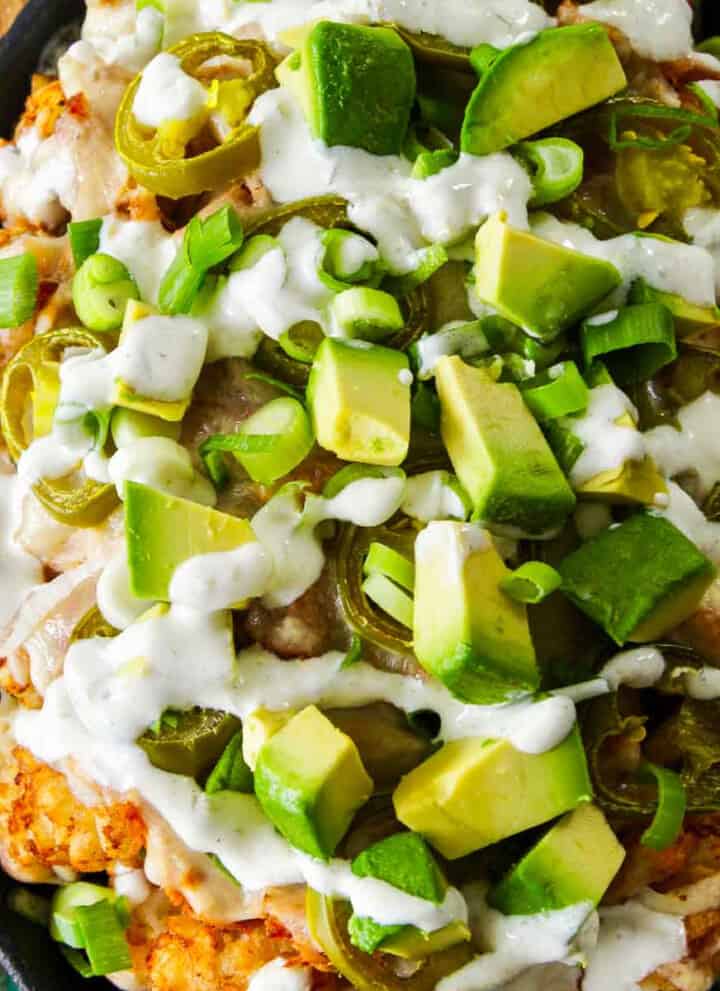 A close up of a oval cast iron pan with tater tots, pulled pork, cut avocado, and drizzled with white sauce.