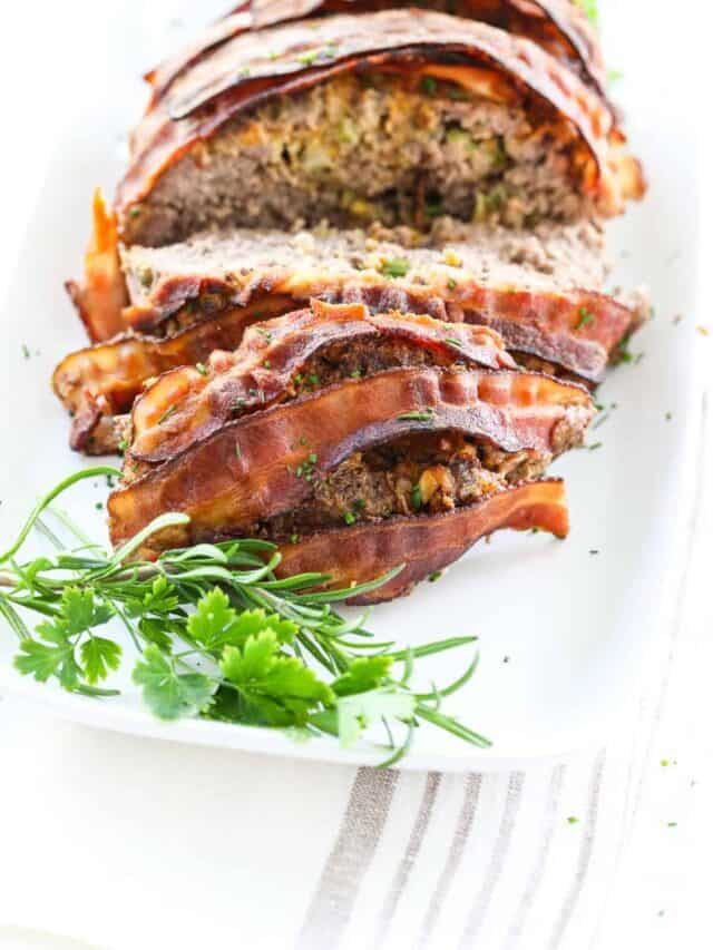 Cooked cheeseburger meatloaf recipe wrapped in bacon on a white platter with herb garnish.