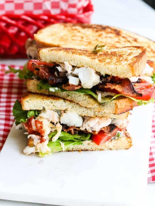 A BLT lobster sandwich cut and stacked on a white cutting board loaded with chunks of lobster and applewood smoked bacon.