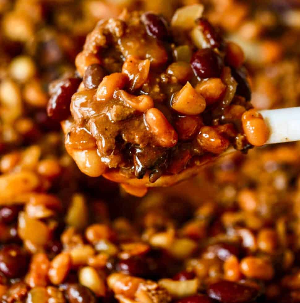 A large white spoon scooping a serving of cowboy baked beans out of a casserole dish.