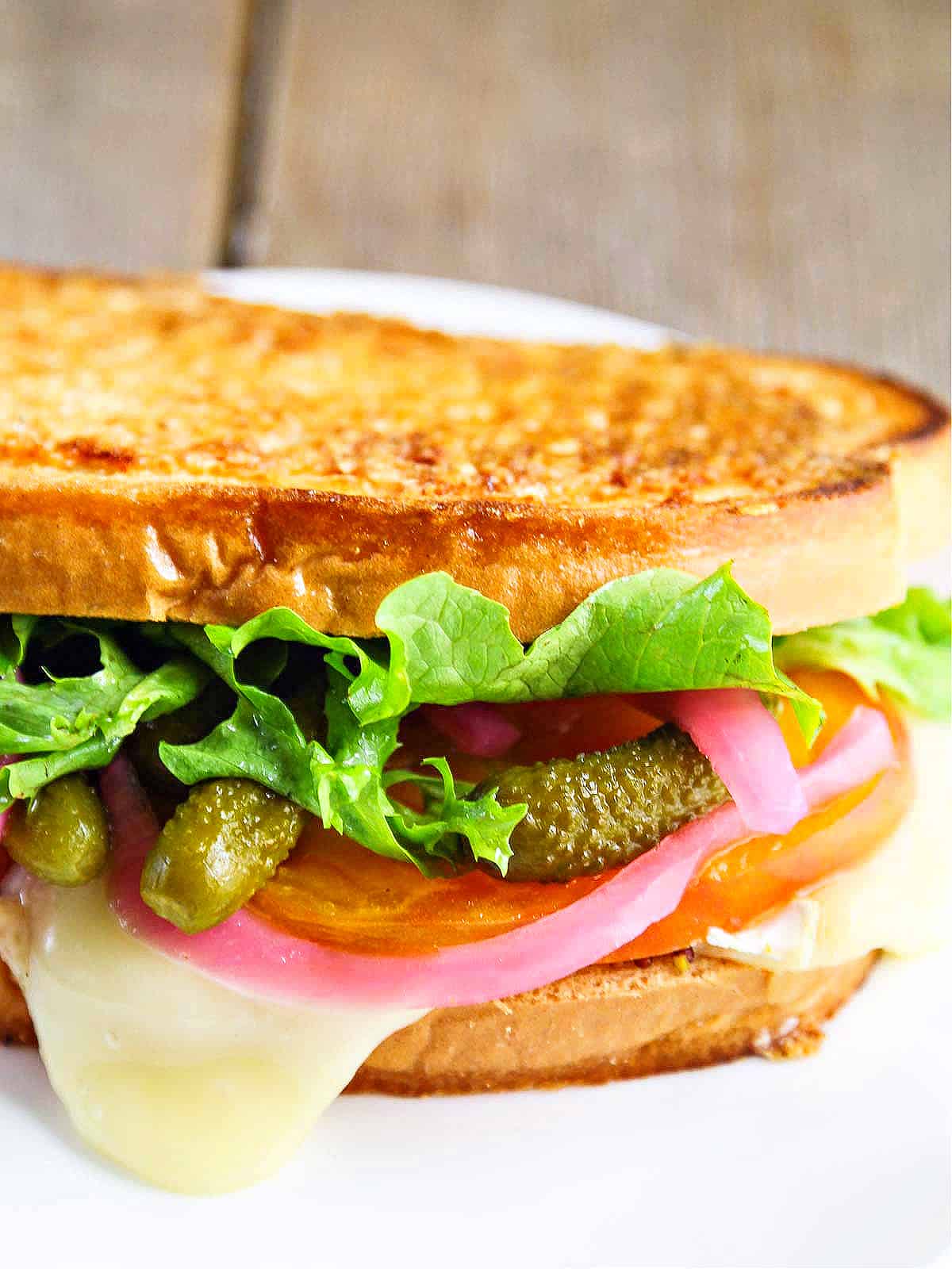 A golden brown grill cheese sandwich stuffed with cheese, cornichon pickles, red pickled onions, and lettuce.