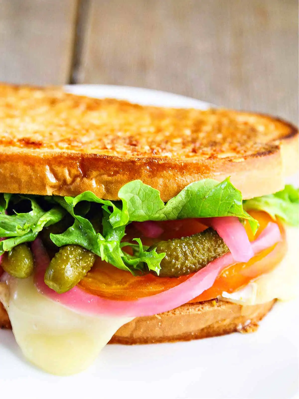 A golden brown grill cheese sandwich stuffed with cheese, cornichon pickles, red pickled onions, and lettuce.