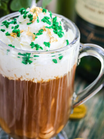 A glass mug with Irish Coffee on a vintage green table topped with green sprinkles and edible gold.