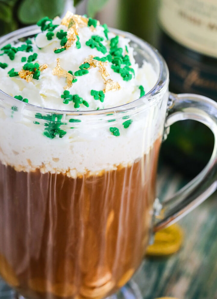A glass mug with Irish Coffee on a vintage green table topped with green sprinkles and edible gold.