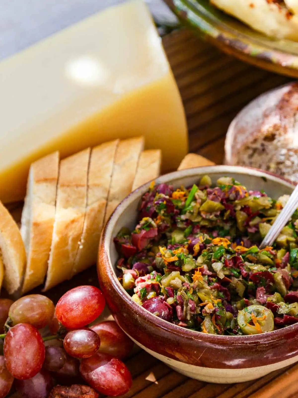 A rustic ceramic bowl filled with vegan olive tapenade, by red grapes and crostini.