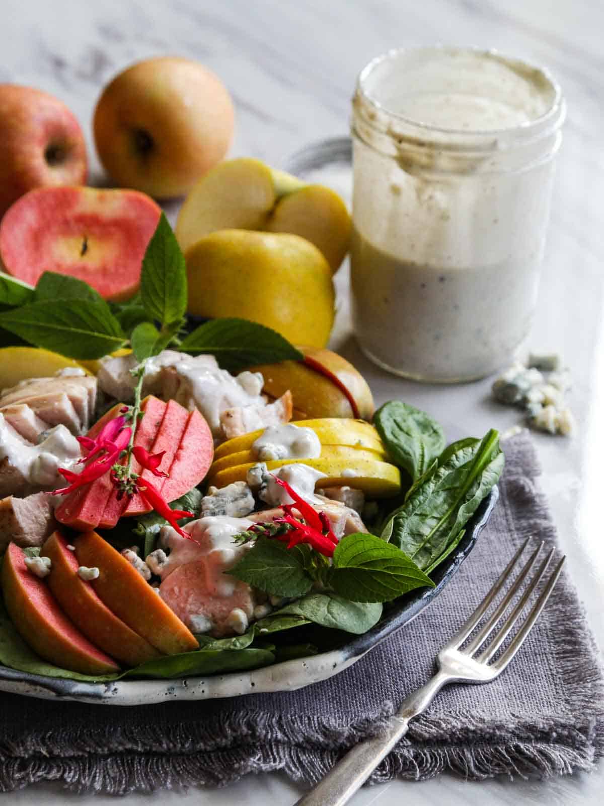 An open jar of blue cheese dressing next to apples and a plate of sliced chicken salad.