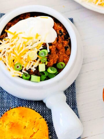 A bowl of turkey chili topped with sour cream, grated cheese, and green onions with corn bread muffins.