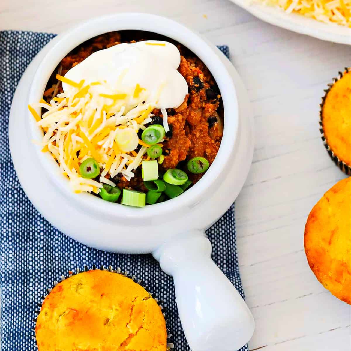 A bowl of turkey chili topped with sour cream, grated cheese, and green onions with corn bread muffins.