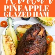 A baked and glazed ham on a white serving platter with pineapple halves and garnish.