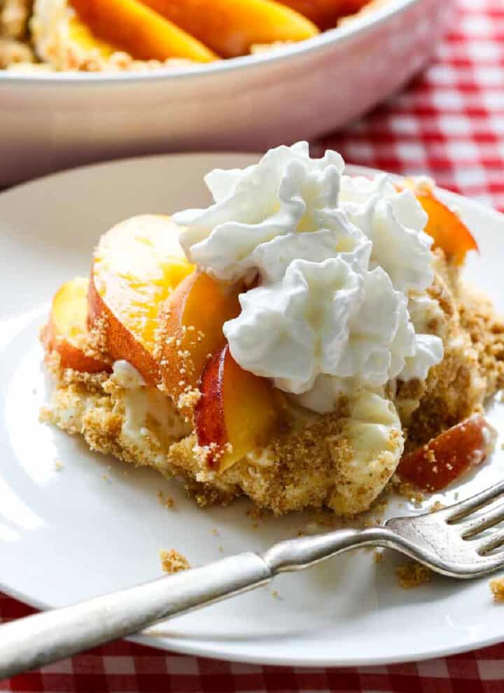 A slice of peach pie on a white plate with a silver fork on a red white check tablecloth.