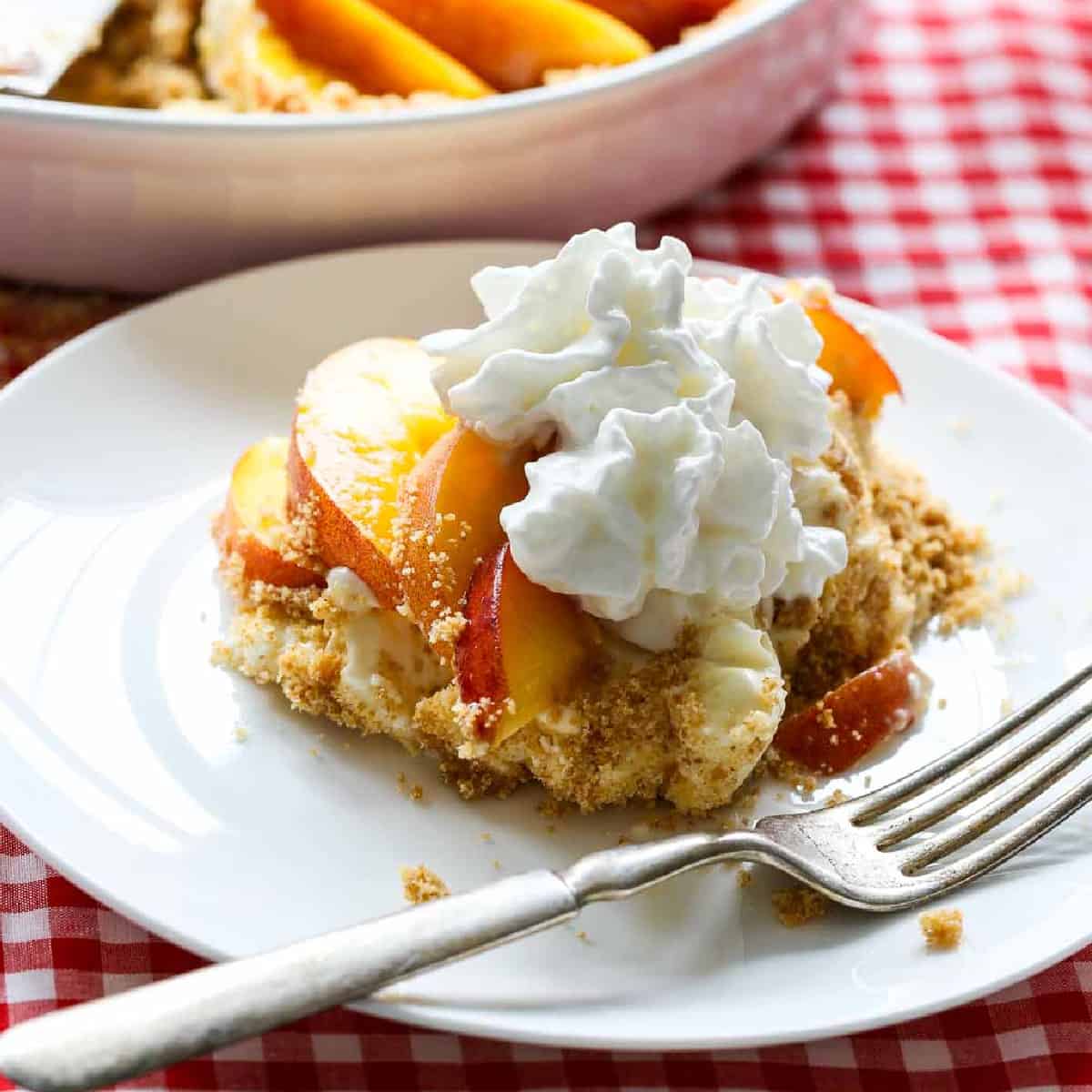 A slice of peach pie on a white plate with a silver fork on a red white check tablecloth.