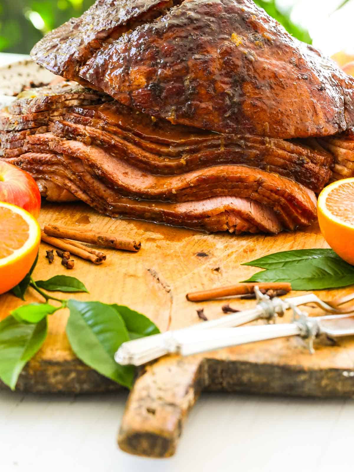Close up of smoked ham on a cutting board garnished with oranges and apples on a cutting board.