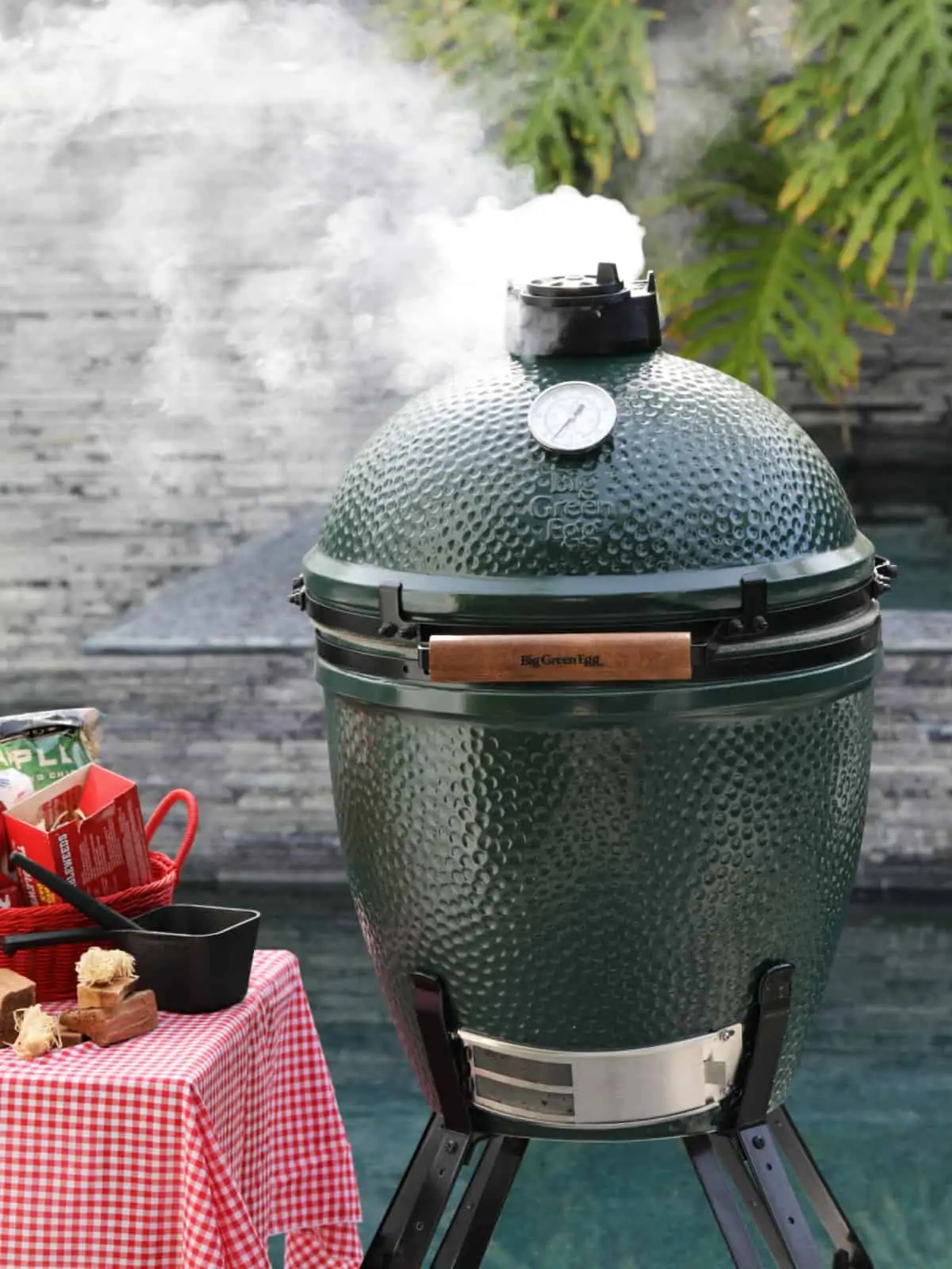 Smoke coming out of the top of a Big Green Egg smoker by a pool.